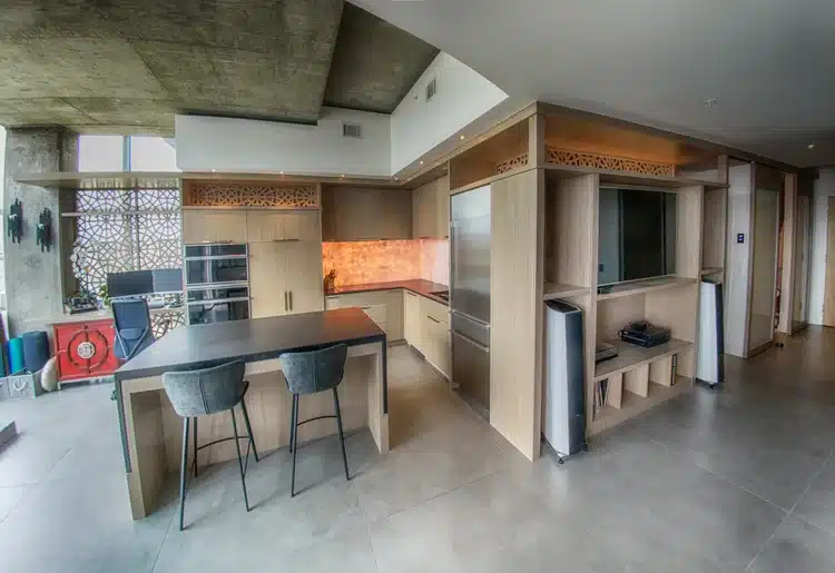 A modern kitchen featuring a minimalist design with light wood cabinets and a large island. Two gray bar stools are positioned at the island. The space has large windows allowing natural light and a view outside, emphasizing open-concept living.